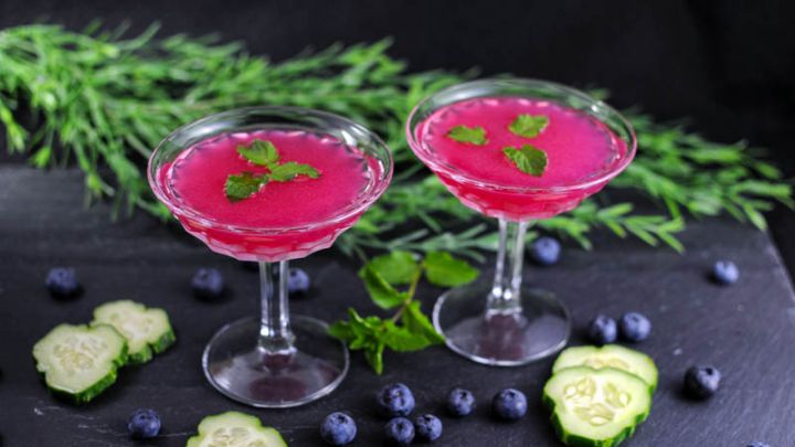 Blueberry Cucumber Gimlet - 2 pink cocktails in coupes with mint garnish. Greenery, blueberries, cucumber slices and mint on slate board.