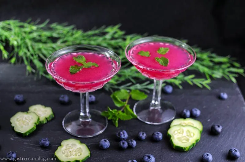 Blueberry Cucumber Gimlet - pink cocktail in 2 coupes with mint leaves on top. Mint, blueberries and cucumbers with greenery on a slate