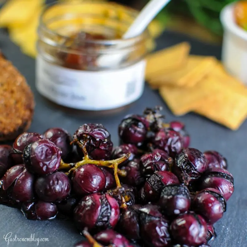 Roasted grapes on the stem on a slate board, cheese, jar of jam and crackers behind