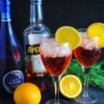 orange Aperol Spritz Cocktails in wine glasses with orange slices. Cut orange and greenery in background with Aperol bottle and blue prosecco bottle
