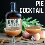 Peach Pie Cocktail - 2 brown cocktails in coupes, cinnamon sticks, peaches, whiskey bottle and greenery in background