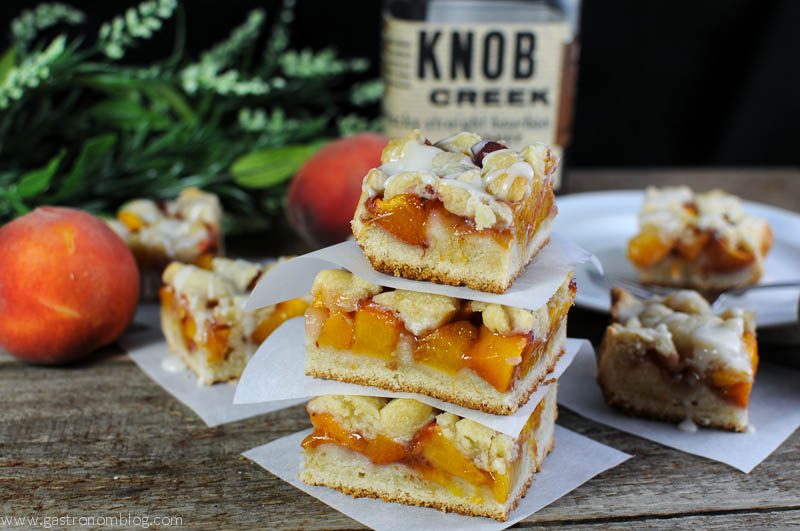 Peach Shortbread bars stacked, some in background on plate. Peaches and bourbon bottle in background