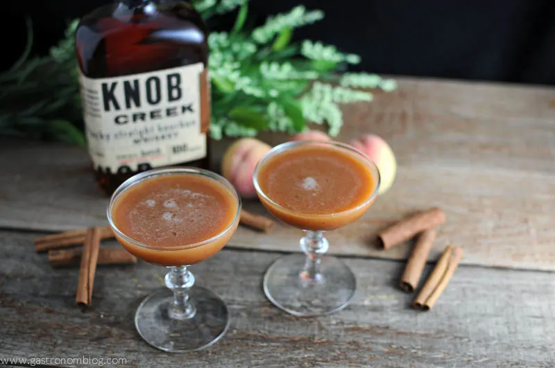 Peach pie cocktails in two coupes with cinnamon sticks, peaches and bourbon bottle in the background