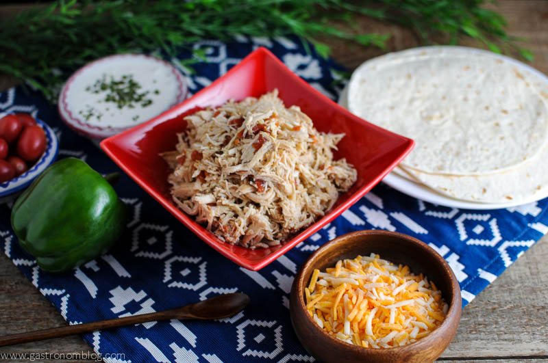 Shredded chicken tacos in a red bowl surrounded by bowls of cheese, sour cream and tomatoes, and tortillas. All on a blue and white napkin