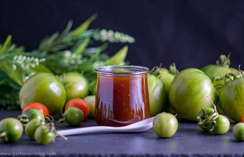 Green tomato jam in jar with white spoon. Green tomatoes and flowers all around