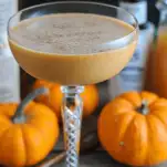 Pumpkin cocktail in a coupe with pumpkins and bottles behind
