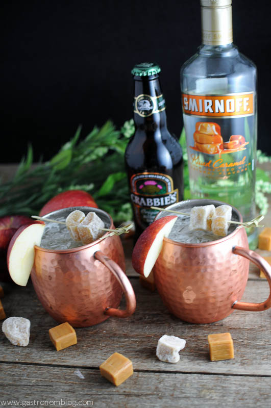 Moscow Mules in copper cups with apple slices and ginger. Caramel vodka and ginger beer bottles behind, with chunks of caramel candies on wood table