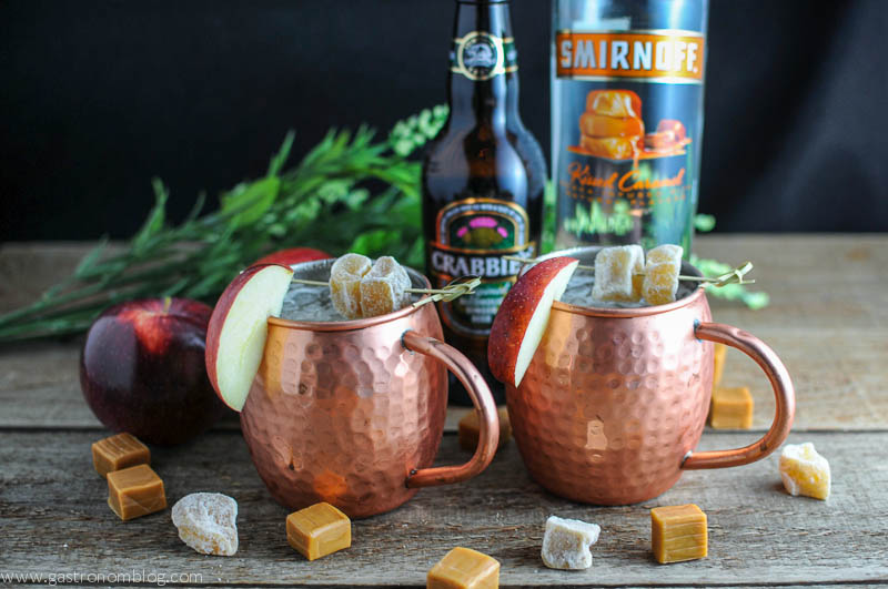 Copper Moscow Mule Mugs with caramel apple mule, crystallized ginger on cocktail picks, caramel squares and apples. Ginger beer bottle and caramel vodka bottle in the background