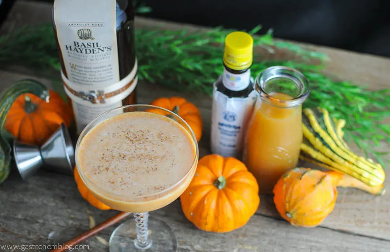 Pumpkin Harvest Cocktail in a coupe. Pumpkins behind with bottles