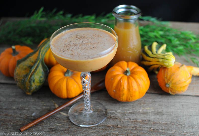 Pumpkin Harvest Cocktail - orange creamy cocktail in coupe with pumpkins and gourds, wooden spoon and jar of apple cider behind
