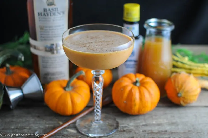 Pumpkin Harvest cocktail in cocktail coupe with mini pumpkins and jiggers, whiskey bottle and bitters bottle in background