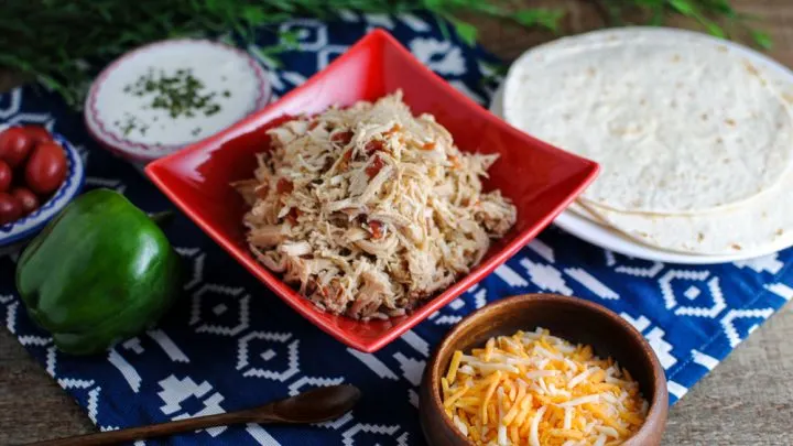 Shredded Chicken Tacos in a red bowl with bowls of sour cream, cheese and tortillas around it.