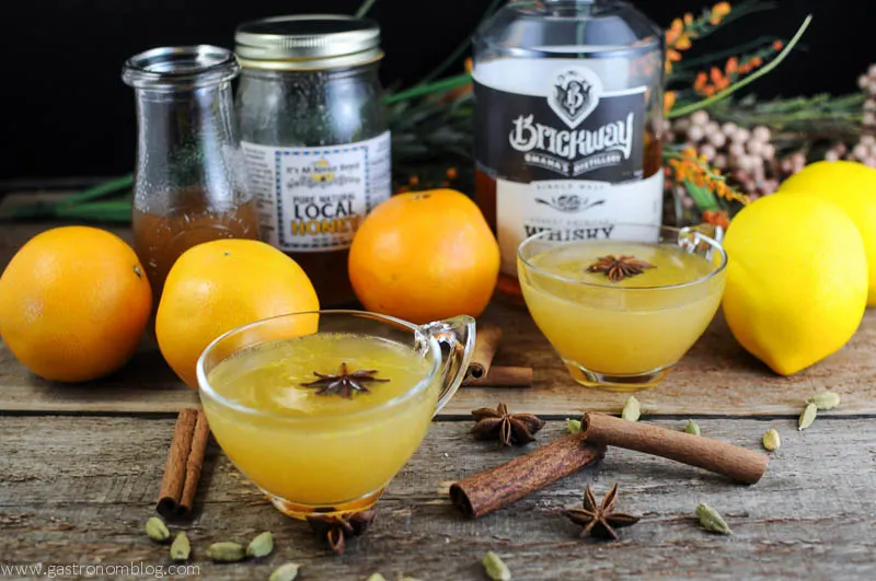 Yellow whiskey punch recipe in punch cups with star anise. Bottles, spices and oranges behind.