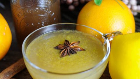 Yellow whiskey punch recipe in punch cups with star anise. Spices and oranges behind with flowers.
