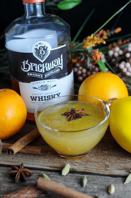 Yellow whiskey punch recipe in punch cups with star anise. Bottles, spices and oranges behind.