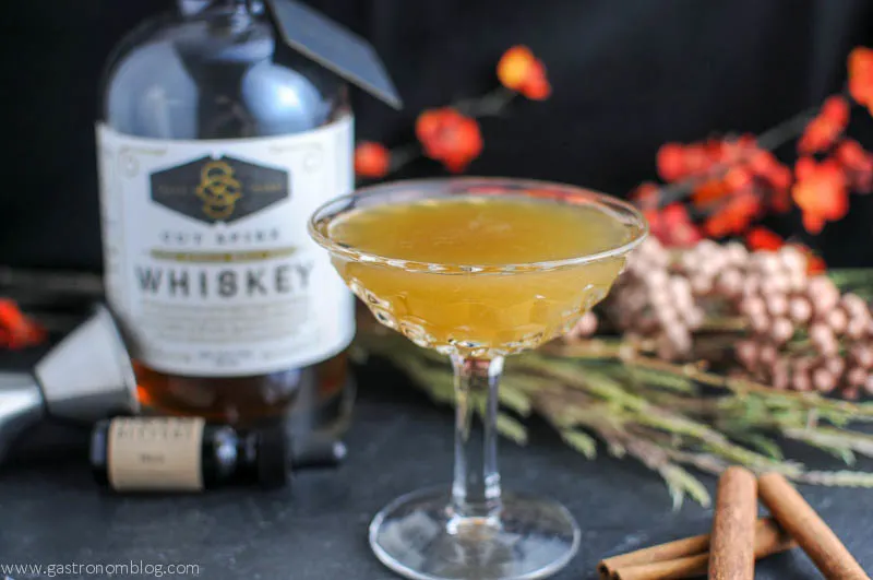Gold cocktail in coupe, whiskey and bitters bottles in background with cinnamon sticks and flowers