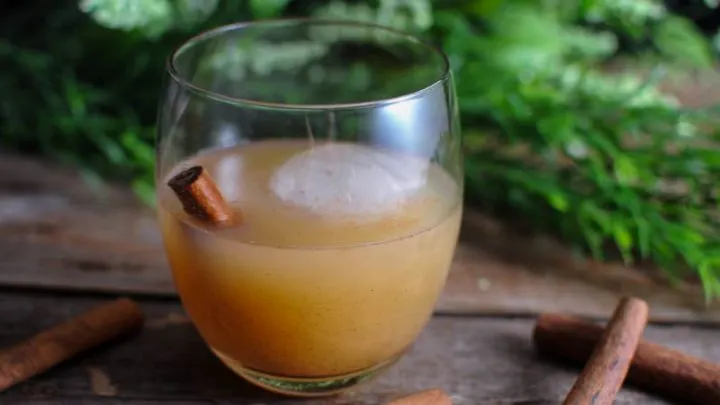 Spiced Pear Tequila Cocktail in wine glass - tan cocktail with ice ball. Cinnamon stick in glass and on wood table.