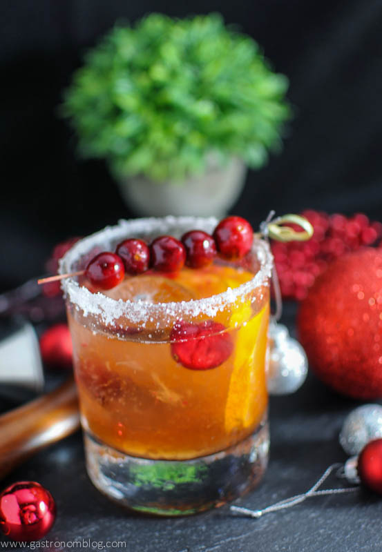 Christmas Old Fashioned - gold cocktail in rocks glass with sugar rim, cranberries on pick. Christmas ornaments and greenery behind
