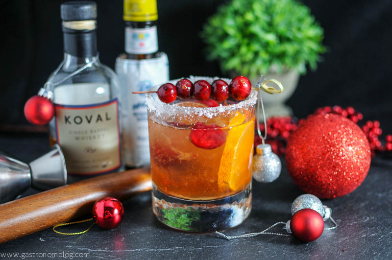 Christmas Old Fashioned - golden cocktail in rocks glass with sugar rim and cranberries on pick. Muddler, bottles and Christmas ornaments in background