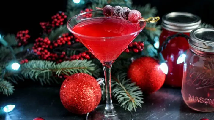 Red cocktail in martini glass with cranberries on pick. Evergreen and Christmas ornaments in background with red syrup filled jars
