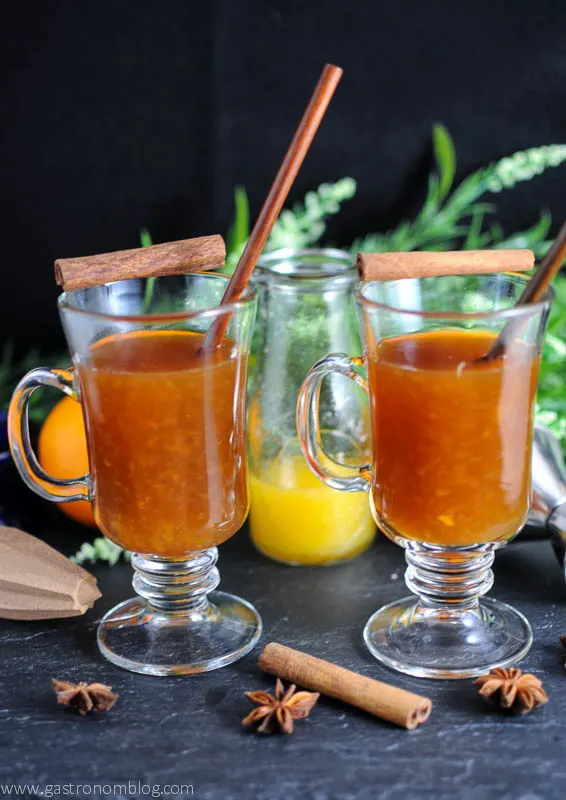 Brown hot toddy in glass mugs with wooden spoon and cinnamon sticks. Orange juice and flowers in background.