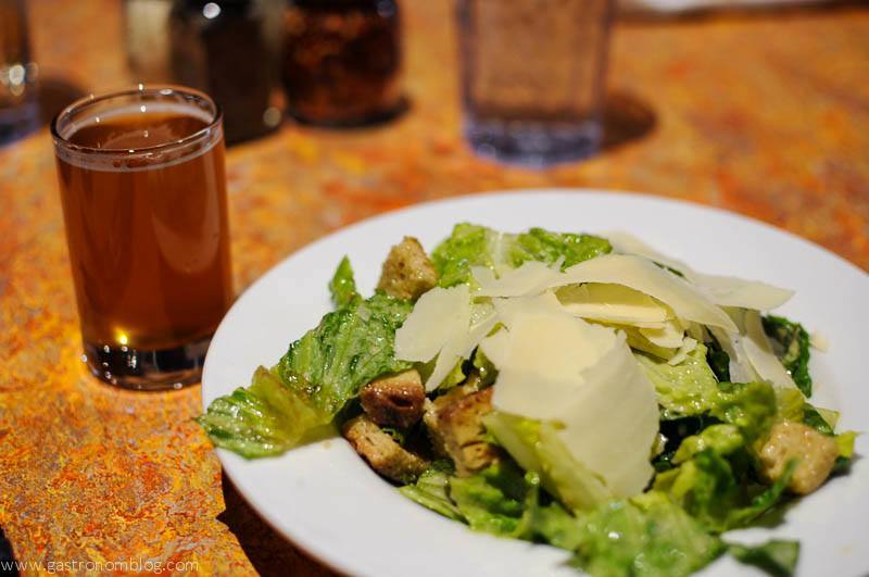 Salad in white bowl with parmesan slices on top. Beer glass on left side. 
