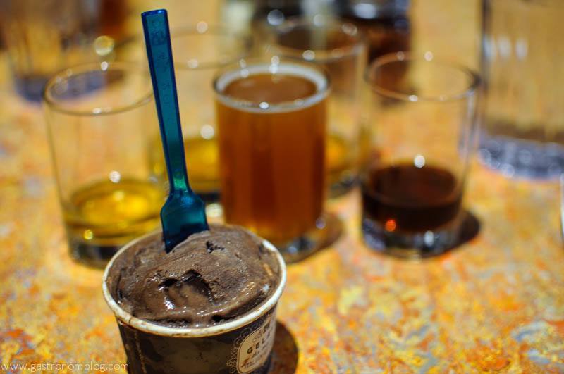 Chocolate gelato in paper cup with blue plastic spoon. Beer glasses behind - full brown beer in middle glass. 