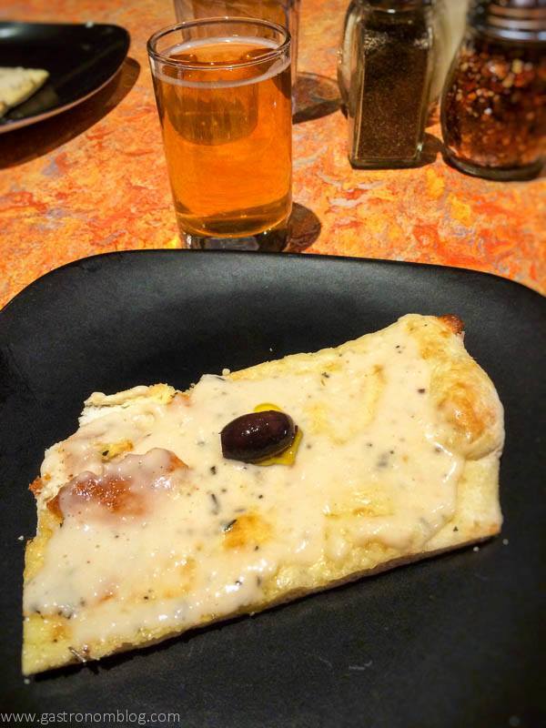 Slice of pizza with olive on it, on a black plate. Beer glass in background