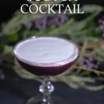 Purple cocktail in coupe with white cream layer on top. Purple flowers in background
