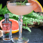 Pink cocktail in coupe with grapefruit slice, rosemary sprig. Gin bottle, grapefruit half and flowers in background