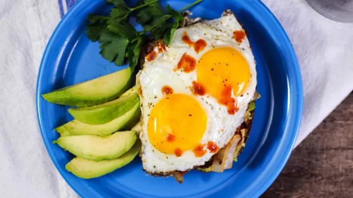 Top shot of egg breakfast sandwich on blue plate. Sliced avocado next to sandwich
