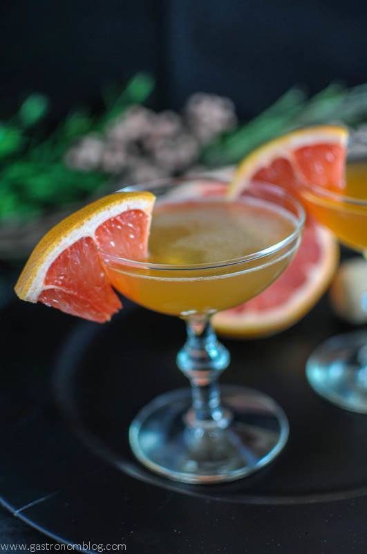 Gold cocktail in coupe with grapefruit slice on black plate. Flowers in background