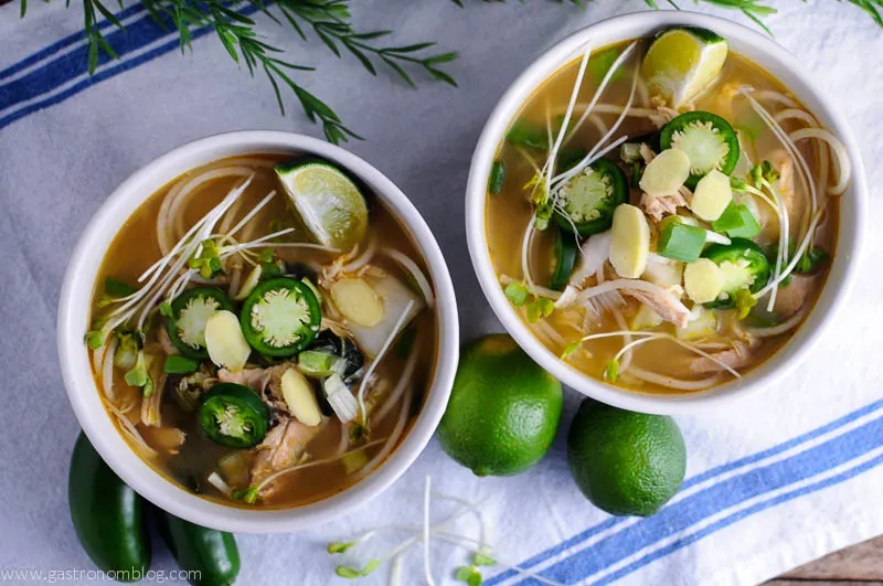 Slow Cooker Chicken Pho soup in white bowls topped with lime wedges, jalapeno slices, and more, limes and jalapenos next to bowls on white napkin