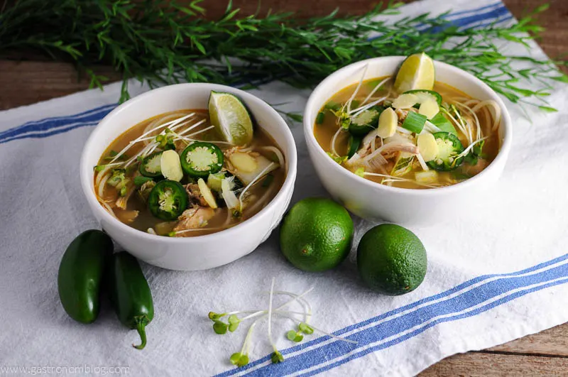 Slow Cooker Chicken Pho soup in white bowls topped with lime wedges, jalapeno slices, and more, limes and jalapenos next to bowls on white napkin