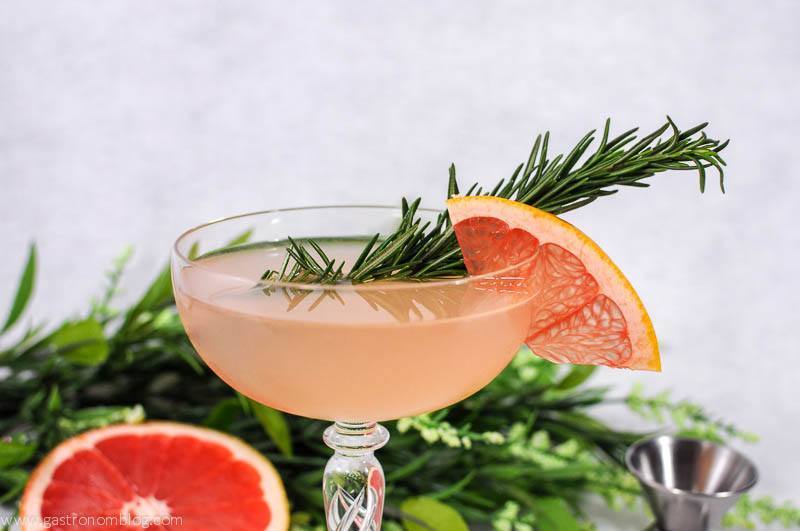 Pink cocktail in coupe with grapefruit slice and rosemary sprig. Grapefruit and flowers in background