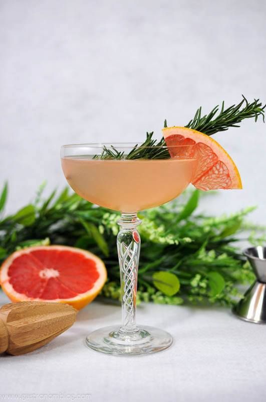 The French Tart Cocktail - pink cocktail in coupe with grapefruit slice and rosemary sprig. Half grapefruit, greenery, reamer and jigger in background.
