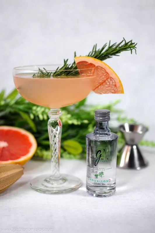 French Tart cocktail in coupe with grapefruit wheel and rosemary sprig. Grapefruit and gin bottle with flowers in the background