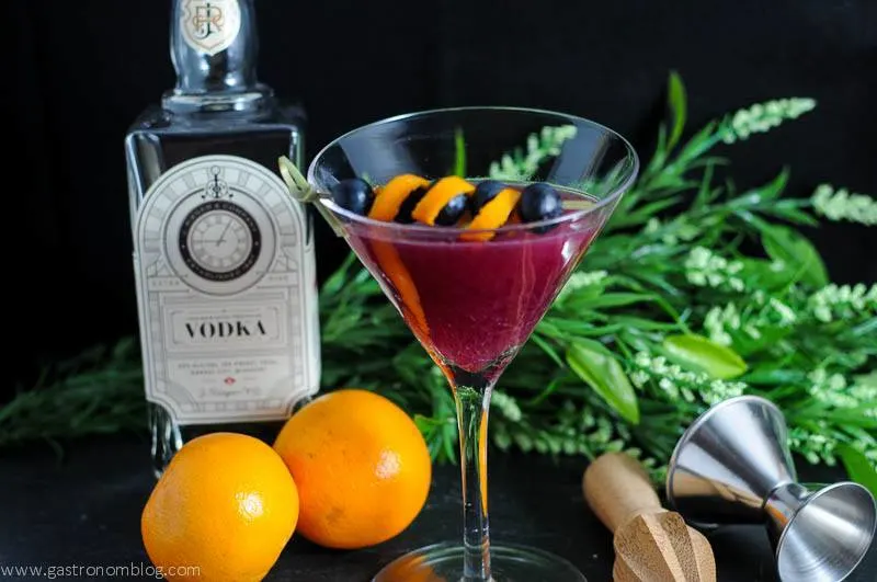 Garnet cocktail in a martini glass with oranges, jigger, reamer and vodka bottle in the background