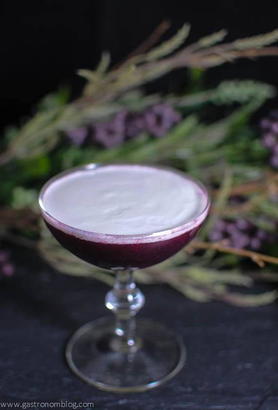 Purple cocktail topped with white cream in a coupe. Flowers behind.