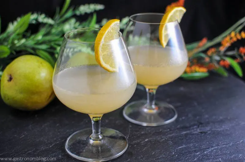 Truffled Anjou cocktail in two brandy snifters with lemon wheel garnishes, pear and flowers in background