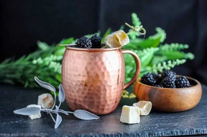 Blackberry Sage Kentucky Mule in a copper mug, wooden bowl of blackberries, greenery in background