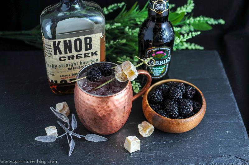 Blackberry Sage Mule in a copper mug, bourbon bottle, ginger beer, blackberries and greenery in background