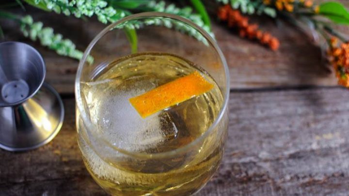 Gold cocktail in glass with large ice cube and orange peel. Jigger and flowers behind glass on wood table