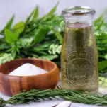 Green Rosemary simple syrup in a jar, wood bowl of sugar, green floral behind
