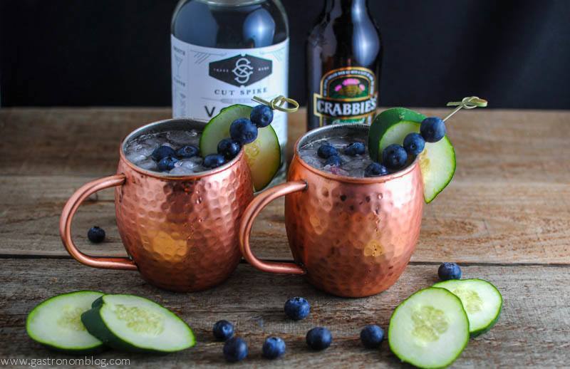 Blueberry Cucumber Moscow Mule in copper mugs, blueberries around and on cocktail picks. Cucumber slices on table and on cups. Vodka bottle and ginger beer bottle behind.