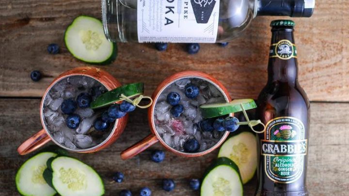 Top shot of mules in copper cups with cucumber slices and blueberries, vodka and ginger beer bottles on wood table