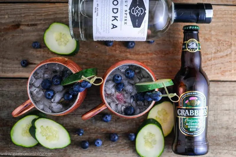 Blueberry Cucumber Moscow Mule top shot on wooden table with blueberries scattered and on cocktail picks. Cucumber slices, vodka bottle and ginger beer bottle.