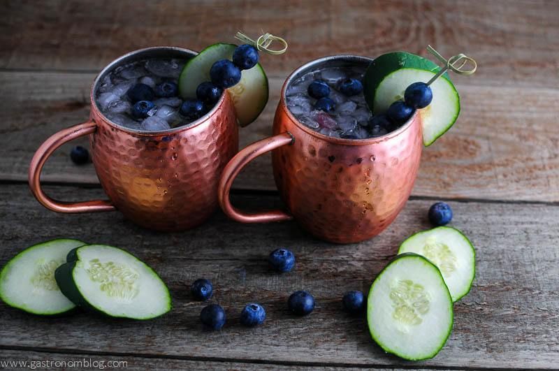 Blueberry Cucumber Moscow Mule in copper cups with blueberries on cocktail pick and cuucmber slices. All on wooden table.