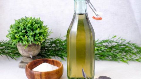 Sage Simple Syrup - green syrup in bottle with wooden spoon and bowl with sugar, sage leaves by bottle. Greenery behind.