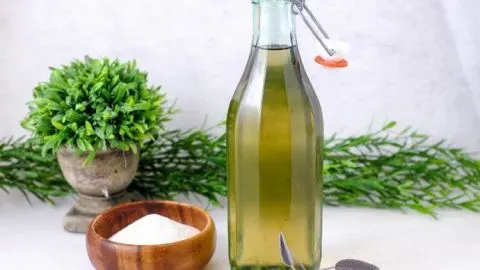 green sage simple syrup in a tall bottle, wood bowl and spoon with sugar. Sage leaves in front and behind bottle